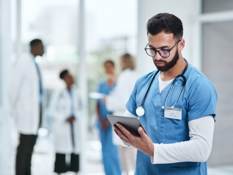 A male doctor standing in a hallway reading something on a tablet, with a group of doctors in discussion behind him.
