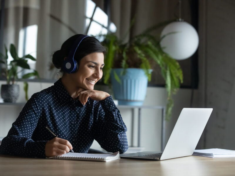 A woman wearing headphones and looking at a computer screen while she takes notes.