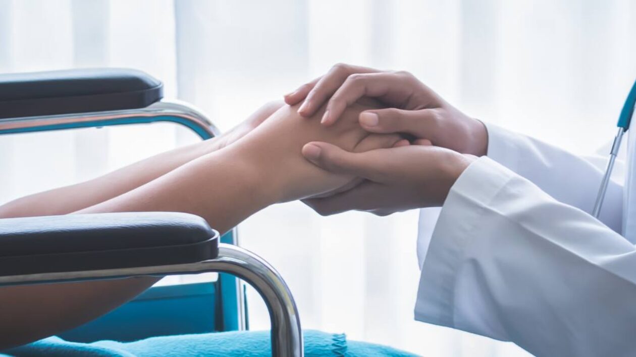 A close up of a doctor in a white coat with a stethoscope around her neck crouching down to a young patient in a wheelchair holding hands.
