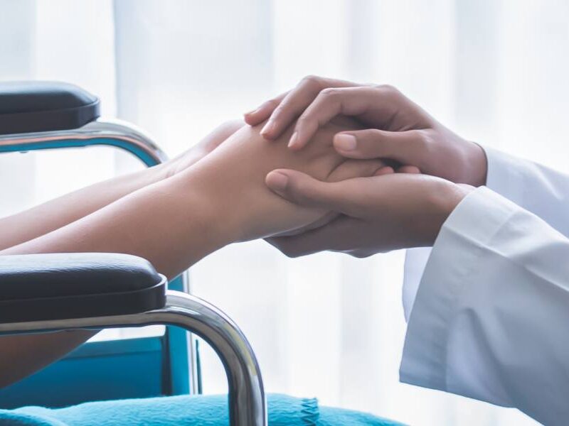 A close up of a doctor in a white coat with a stethoscope around her neck crouching down to a young patient in a wheelchair holding hands.