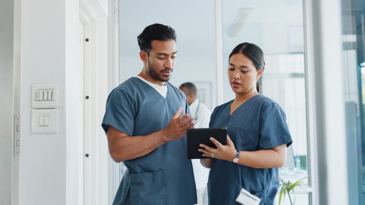 A male nurseand female nurse conversing in a hospital corridor whilst holding a tablet.