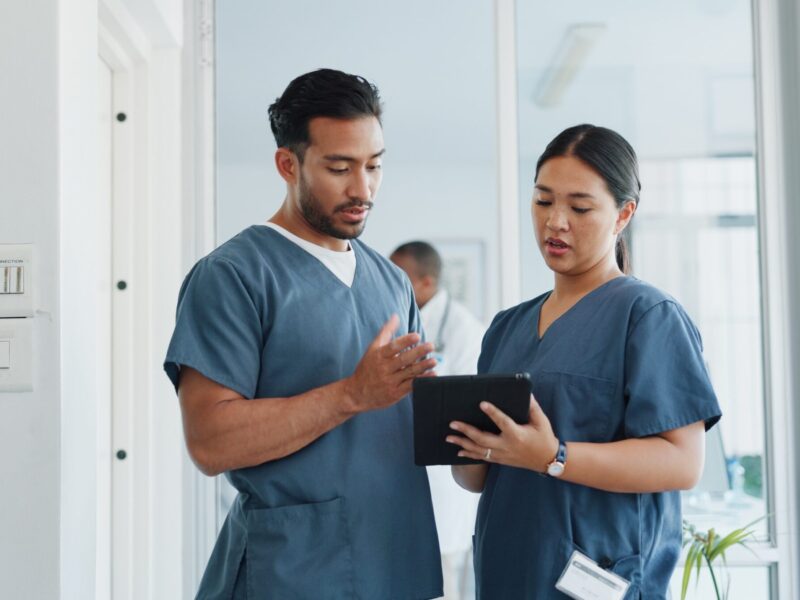 A male nurseand female nurse conversing in a hospital corridor whilst holding a tablet.